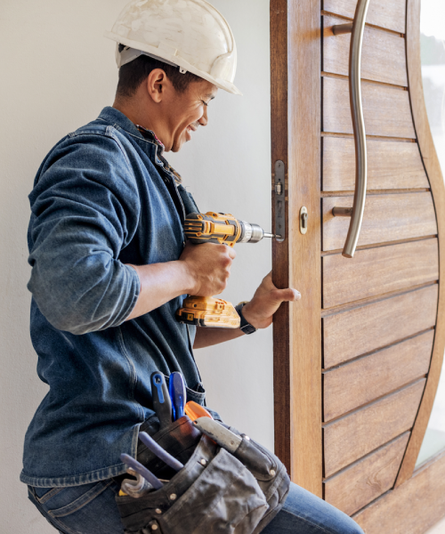 A worker wearing a hard hat and tool belt uses a power drill to install or repair a lock